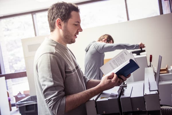 Male students looks over project while another student works on the printing press